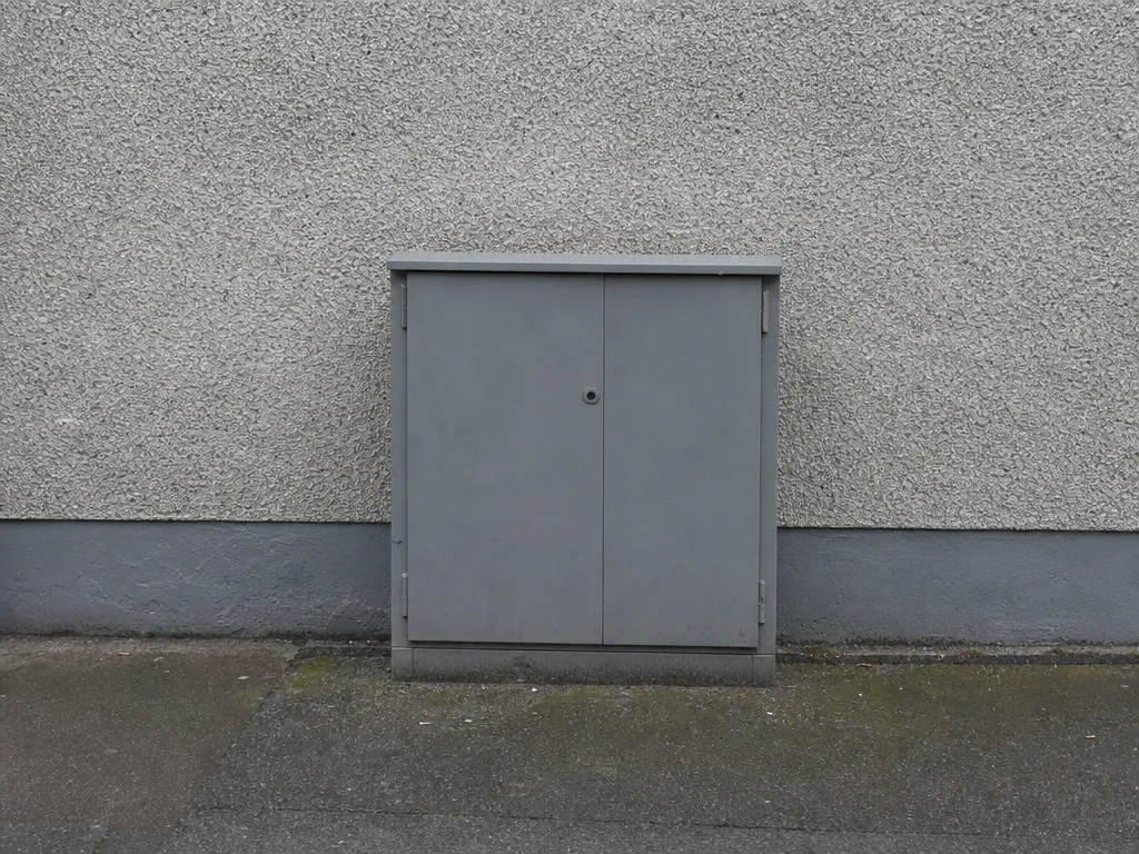 A plain grey utility cabinet standing against a blank pebbledash wall on an ordinary pavement.