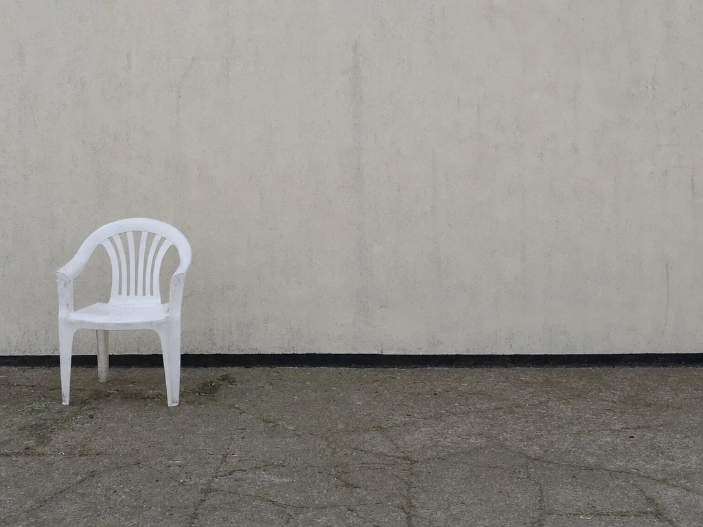 A single white plastic garden chair standing off-centre on a cracked concrete forecourt in front of a plain wall.