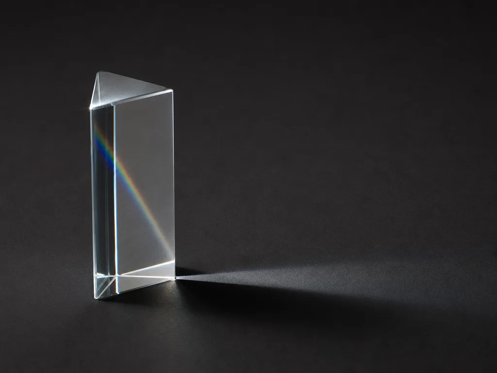 A clear glass prism on a graphite surface under hard cool directional light, splitting a faint spectrum, set off-centre to the left.