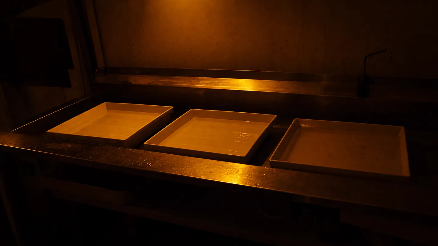 Three shallow developing trays set in a long sink under a dim safelight, still liquid catching the light.