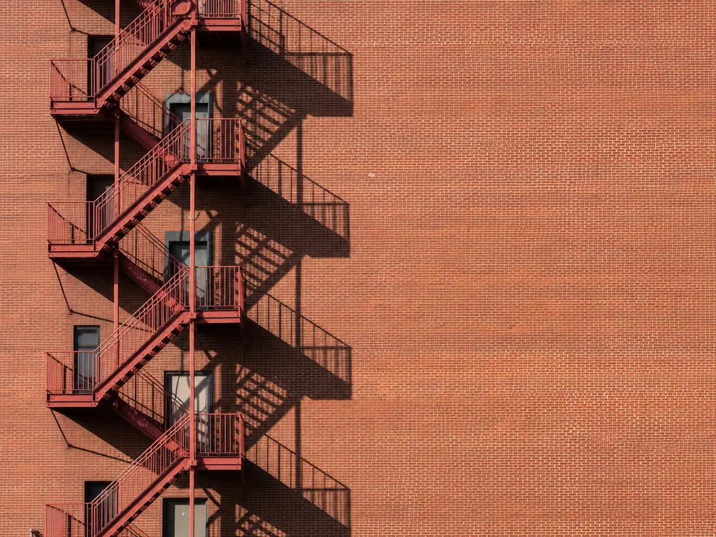 A red iron fire escape zig-zagging up the left of a sunlit brick wall — the source photograph.