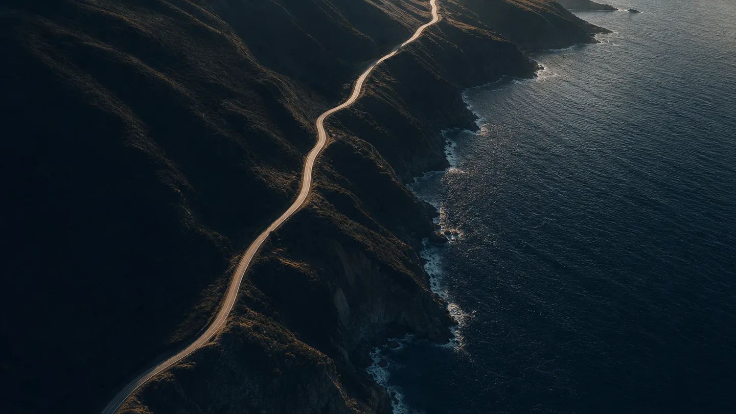 A high aerial of a lone road curving between dark cliffs and open sea in hard low afternoon light.