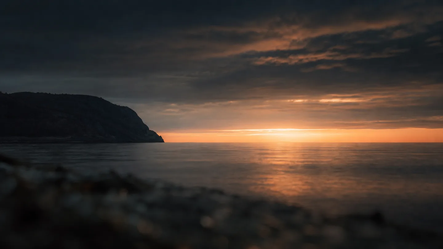 A coastal horizon at last light, long tonal gradients from deep shadow to a single bright band on the water.